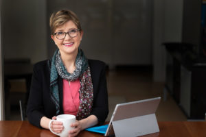 Woman looking at you in front of her computer holding a mug.