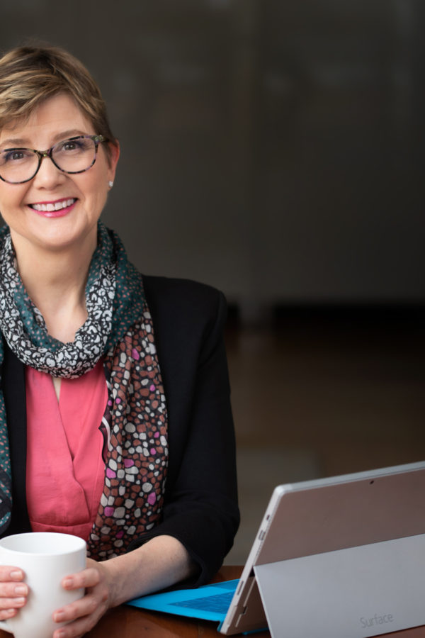 Woman looking at you in front of her computer holding a mug.